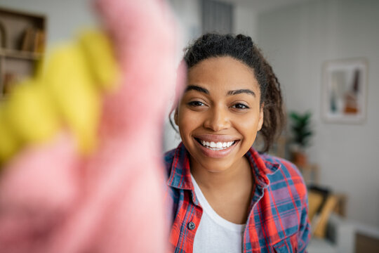 Cheerful Young Black Woman In Rubber Gloves Washes Mirror With Rag, Enjoys Cleaning In Living Room Interior