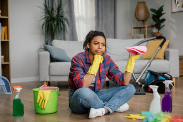 Sad bored young black woman in rubber gloves sits on floor with cleaning supplies, suffers from a...