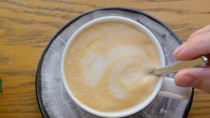 Top view of a cup with cappuccino, milky coffee foam is stirred with a spoon by woman. Tasty and invigorating drink in the morning allows you to wake up. On the table is a mug of coffee with milk foam
