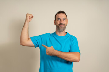 Handsome funny bearded hispanic man pointing at his biceps and smiling over beige background