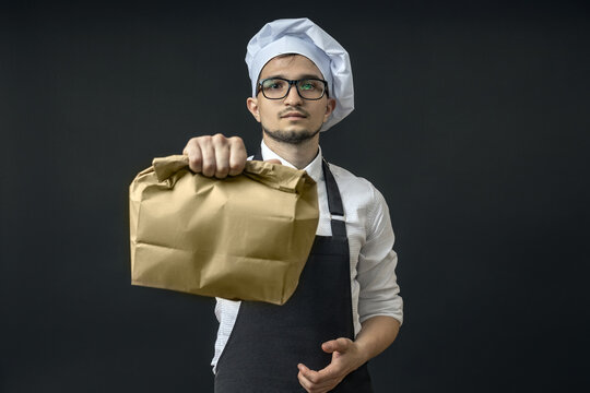 a young serious chef holds out a paper bag to the camera on a black background.