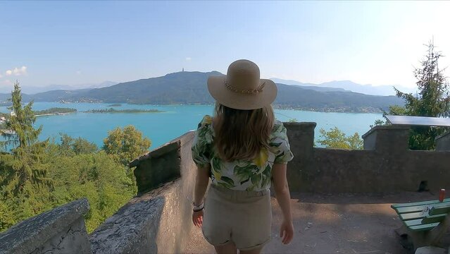 Tourist woman with summer hat enjoying panoramic view on alpine lake Lake Woerth (Woerthersee) in summer seen from platform Hohe Gloriette, Poertschach, Carinthia, Austria, Europe. Karavanks mountains