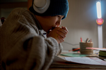 A teenager does his homework in a cold apartment. He sits at a table wearing a hat and a warm...