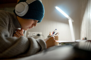 A teenager does his homework in a cold apartment. He sits at a table wearing a hat and a warm sweater. Energy crisis, there is no heating in the house