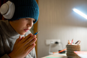 A teenager does his homework in a cold apartment. He sits at a table wearing a hat and a warm sweater. Energy crisis, there is no heating in the house