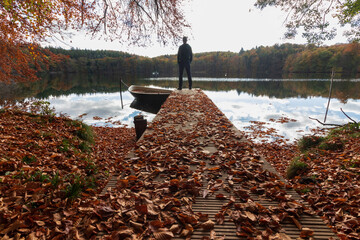 Man standing on a jetty  covered with autumn foliage by the lake. 
