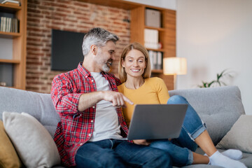 Obraz premium Smiling mature european husband hugs female, pointing finger at computer, sits on sofa in living room