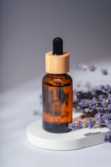 Amber dropper bottle with serum, tonic or essential oil on grey concrete podium. Grey background with lavender flowers.
