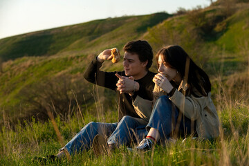 The concept of young people who are happily and having fun on a picnic in nature in the warm season, a young guy with his girlfriend eat ready-made hamburgers in nature with appetite
