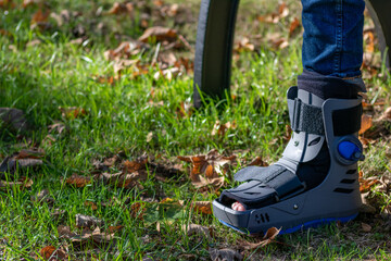 Boy with broken foot and orthopedic shoe or walker after bone fracture rests in public park on...