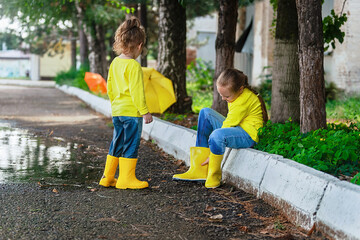 two little cheerful girls in bright yellow jackets and rubber boots after the rain in the warm...