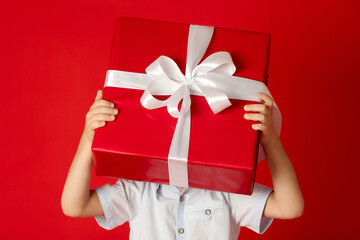 In hands of little boy is large red gift box with white bow, behind which he hid. Boy hid his face...