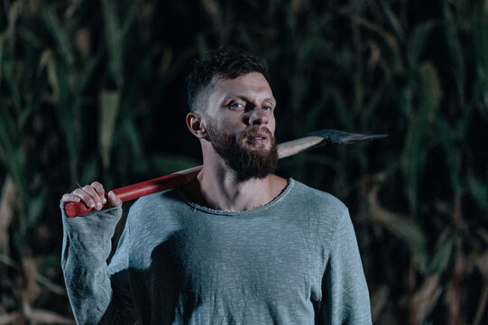 Scary man with ax stands in corn field and looks into camera at night. Close-up portrait. Halloween concept