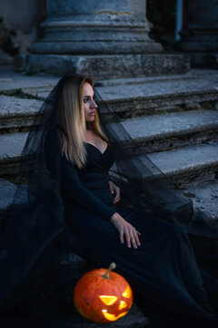 A young woman witch in black clothes sits near the old castle and holds in her hands a luminous pumpkin with a terrible face.