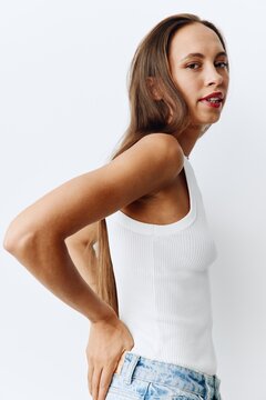 A young woman with beautiful and tanned skin poses fashion Lifestyle in a white tank top and blue jeans against a white wall background