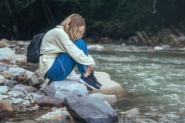 frustrated, tired tourist girl sits alone on the bank of a mountain river on the rocks, she clasped her legs in her hands and laid her head on them during a hike in nature