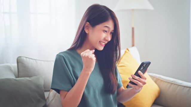 Excited young asian woman hold phone chatting in dating app feel happy sit on sofa at home, Closeup joyful female reading good news on phone in slow motion. Surprised lady celebrating victory on phone