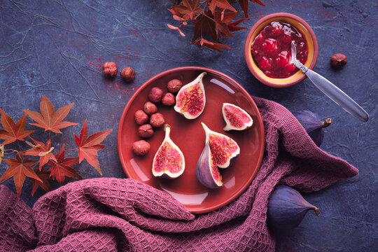 Autumntime background with fresh halved fig fruits on terracotta ceramic plate. Magenta towel and red maple Autumn leaves on dark purple board. Fig jam in ceramic bowl. Natural vibrant Fall colors.