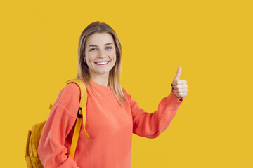Happy caucasian student girl showing ok and excellent sign isolated on bright yellow background. Girl in orange sweatshirt and with backpack shows thumb up looking at camera. Banner.