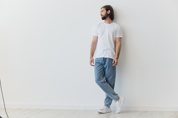 A man with a beard and long hair in a white T-shirt and blue jeans stands against a white wall leaning full-length on it, relaxed style