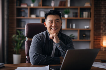 Smiling successful handsome adult korean man at workplace with laptop look at camera