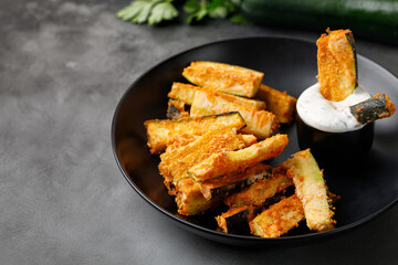 Zucchini sticks baked with cheese, breadcrumbs with cream yogurt sauce. Healthy tasty snack, vegetarian appetizer. Roasted vegetable snack on dark plate.