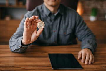 Mature man show bitcoin coin at table with tablet with blank screen in office interior, cropped, close up