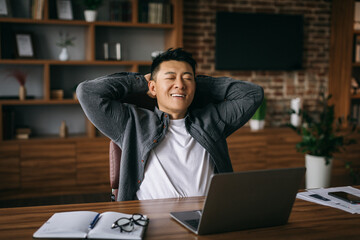 Smiling adult chinese male enjoys rest in chair at table with computer in office interior
