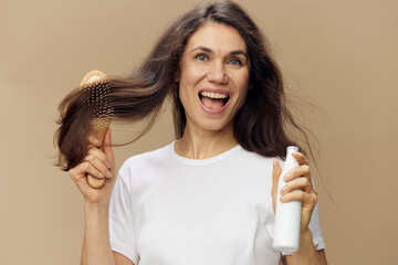 a beautiful, happy, sweet woman stands on a beige background in a white tank top and combs her beautiful, well-groomed, dark, shiny hair with a comb, spraying it with hair product
