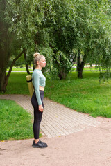 Fototapeta premium A slender woman in sportswear, standing in summer in park, with a green gym mat for sports.