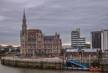 Foto op Canvas Antwerpen, Vlaanderen, België - 10 juli 2022: Langs de Schelde. Beigebruine steen Historisch Loodswezen, scheepvaartloodsen, gevel onder grijs wolkendek. Gedenkteken voor verdwaalde zeelieden en meer © Klodien
