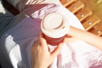 Photography from above of anonymous woman, holding craft paper cup with coffee outside,sunlight on...