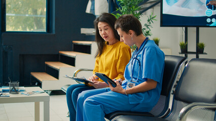 Medical assistant consulting asian woman in hospital lobby, taking notes on digital tablet and filling in report papers. Nurse doing checkup examination with patient in waiting area.