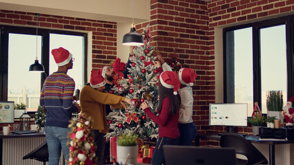 Multiethnic team of people putting christmas tree decor in festive office, preparing for winter season tradition at job. Decorating workplace with xmas ornaments for seasonal holiday.