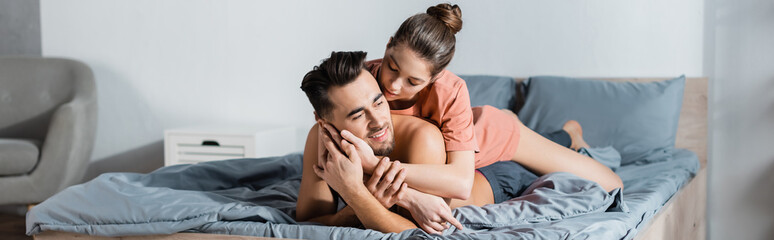 young and hot woman embracing happy boyfriend lying on grey bedding, banner.