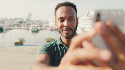 Young man takes selfie on the phone while sitting in harbor with yachts on the background