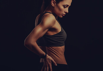 Fototapeta na wymiar Strong sporty woman doing hand massege to relax the tension in low back and sacrum in sport bra top on empty copy space black studio background. Sports exercises injury. Back view.