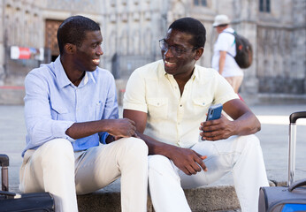 Positive african american travelers friendly talking while sitting with luggage on summer city street.