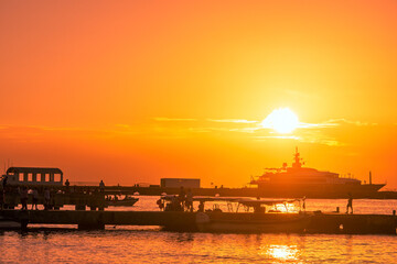 Sunset in the harbor. Ship stand at the sea pier against the backdrop of the setting sun. On the pier are figures of people admiring the sunset.