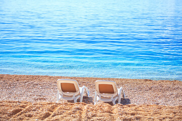  Beach loungers on the seashore . Two chaise-longues on the beach . Coastal vacation concept  - obrazy, fototapety, plakaty