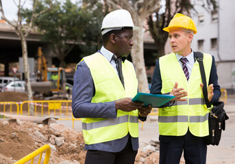 Two men workers working at the laying paving slabs facility discuss the project, holding important...