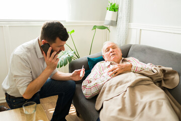 Concerned man taking care of his sick senior father suffering from fever and calling a doctor - obrazy, fototapety, plakaty