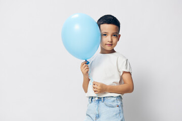 a cute, happy boy stands on a light background in a white t-shirt with a blue balloon in his hand and looks at the camera. Horizontal photo with blank space to insert advertising mockup