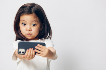 cute, funny, little girl in a white t-shirt stands on a light background with a smartphone in her hand and takes a selfie. Horizontal studio photography with blank space for advertising mockup insert