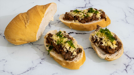 Sliced bread with bruschetta on the side containing red tomato sauce, minced beef, grated smoked Parmesan cheese and green seasoning. Positioned on a white marble background. Mention of finger food.