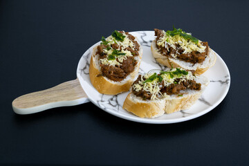 Sliced bread with bruschetta on the side containing red tomato sauce, minced beef, grated smoked Parmesan cheese and green seasoning. Positioned on white ceramic. Mention of finger food.
