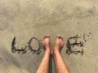 Word “Love” handwritten on beach sand with an unidentified man’s legs replacing and forming the letter V in Love.