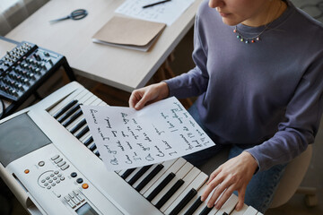 High angle close up of young musician playing synthesizer at home and holding song lyrics sheet,...