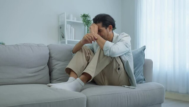 Asian young upset depressed man sitting alone on sofa in living room.