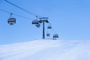 Obraz premium Ski chair lift on top of mountain, ski slope prepared for skiing, minimal style blue monochrome winter in Sheregesh ski resort. White snow, clear sky, cable road and small people on peak
